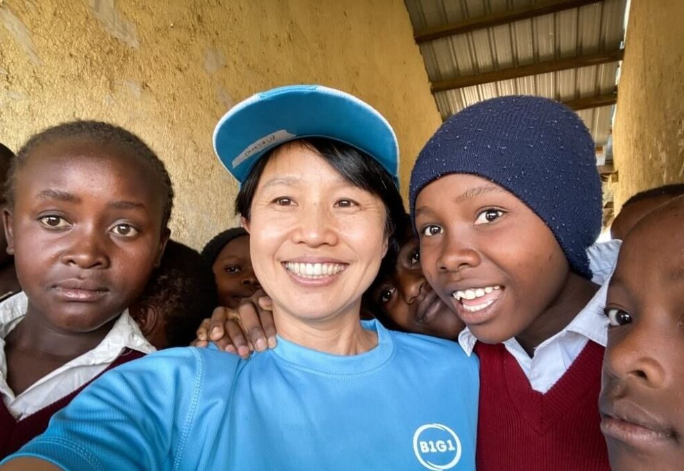 A woman in a blue B1G1 shirt and cap takes a selfie with a group of school children in red uniforms, who gather around her with bright smiles. This image reflects the positive impact of the donations on the local community.