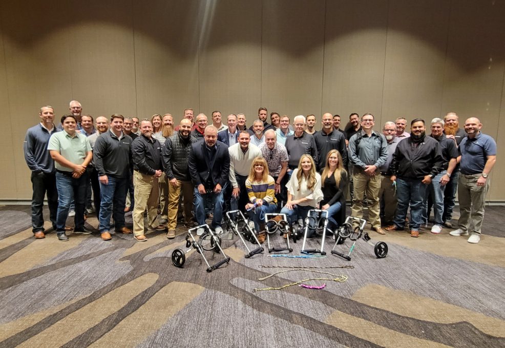 A large group of participants proudly standing together after successfully assembling pet wheelchairs during a team building event, showcasing their completed projects in front of them. The focus is on teamwork and making a meaningful impact on animal welfare.