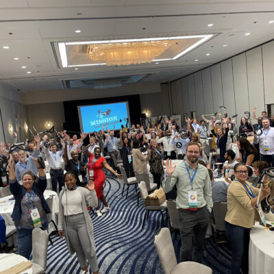 A large group of participants enthusiastically wave during a Mission ImPAWssible team building event, with tables set up for assembling pet wheelchairs in the background. thumbnail