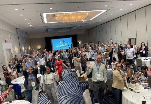A large group of participants enthusiastically wave during a Mission ImPAWssible team building event, with tables set up for assembling pet wheelchairs in the background.