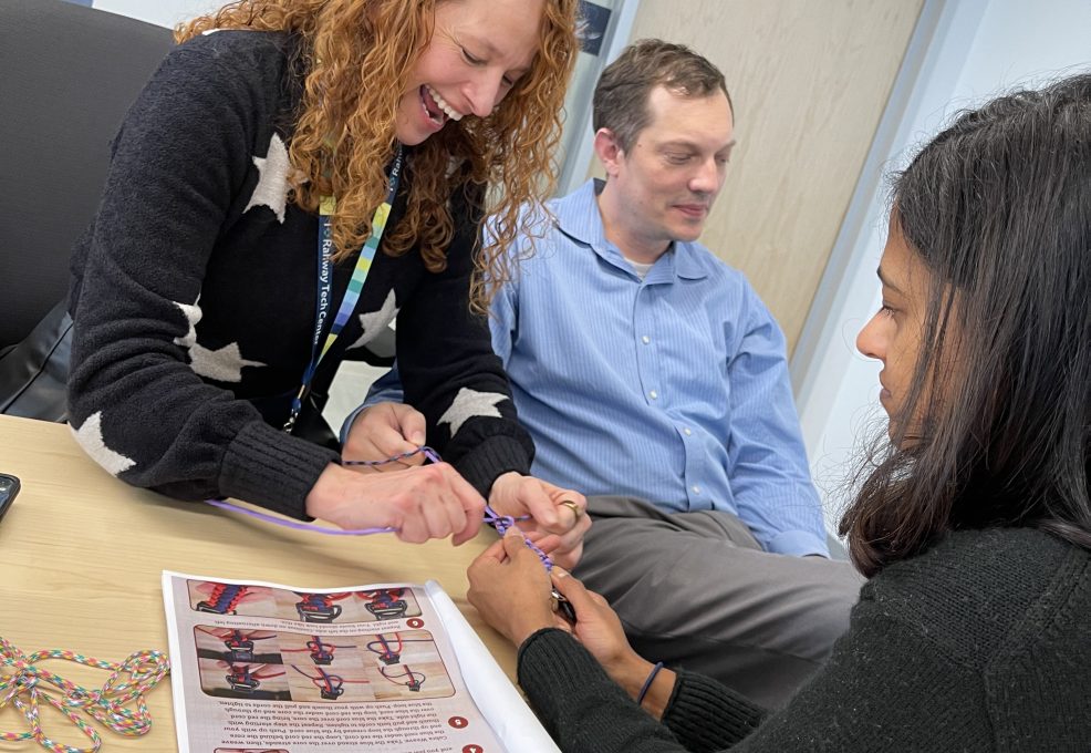 Three team members working together, smiling as they assemble components for a pet wheelchair. They focus on tying ropes while a Mission ImPAWssible brochure is on the table. This scene highlights teamwork and collaboration in creating life-changing mobility aids for pets in need.