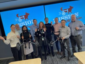 Group of smiling participants posing with assembled pet wheelchairs, standing in front of a screen displaying "Mission ImPAWssible" branding. They are holding various components, ropes, and showing enthusiasm after successfully completing tasks. This event combines teamwork, compassion, and fun while making a real impact for pets in need of mobility.