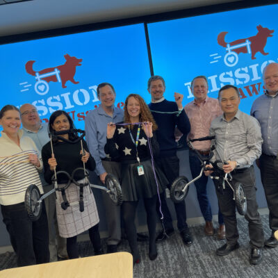 Group of smiling participants posing with assembled pet wheelchairs, standing in front of a screen displaying 