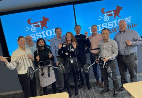 Group of smiling participants posing with assembled pet wheelchairs, standing in front of a screen displaying 