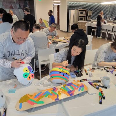 Participants are busy decorating helmets and skateboards during a CSR team building event. The table is filled with various markers and materials as individuals focus on creating colorful and unique designs. thumbnail