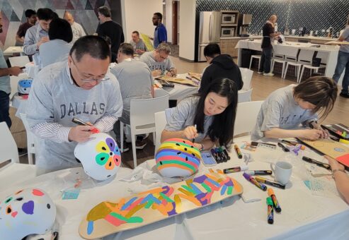 Participants are busy decorating helmets and skateboards during a CSR team building event. The table is filled with various markers and materials as individuals focus on creating colorful and unique designs.