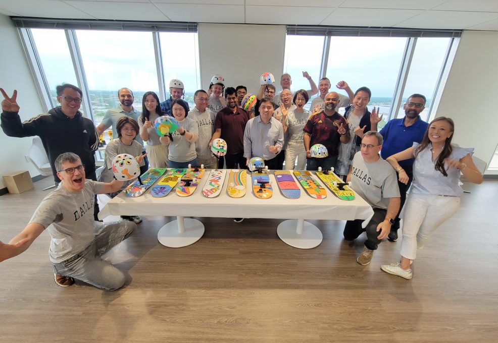 A large group of people celebrating around a table displaying their custom-decorated skateboards and helmets. The skateboards are creatively painted with various colorful designs. Some participants are posing with helmets, and the group looks cheerful, signaling a successful team building event.