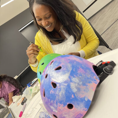 A woman with long black hair, wearing a yellow sweater, is happily painting and decorating a helmet during a team building event. thumbnail