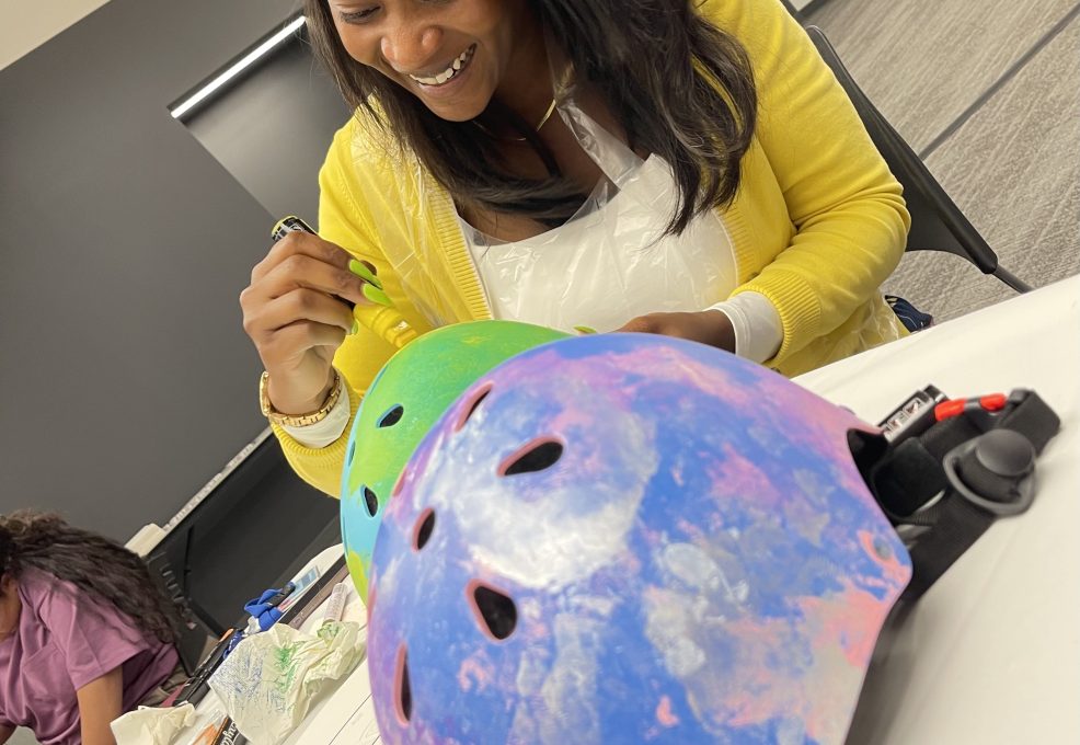 A woman with long black hair, wearing a yellow sweater, is happily painting and decorating a helmet during a team building event.