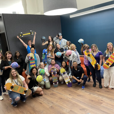 A group of participants proudly displaying their decorated skateboards and helmets after a successful team building event. The group, consisting of around 20 individuals, poses with their custom creations in a brightly lit modern office space. The skateboards and helmets are painted in various colorful designs, showcasing the team's creativity and collaboration. The event celebrates teamwork and artistic expression, bringing everyone together for a charitable cause. thumbnail