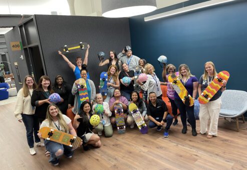A group of participants proudly displaying their decorated skateboards and helmets after a successful team building event. The group, consisting of around 20 individuals, poses with their custom creations in a brightly lit modern office space. The skateboards and helmets are painted in various colorful designs, showcasing the team's creativity and collaboration. The event celebrates teamwork and artistic expression, bringing everyone together for a charitable cause.