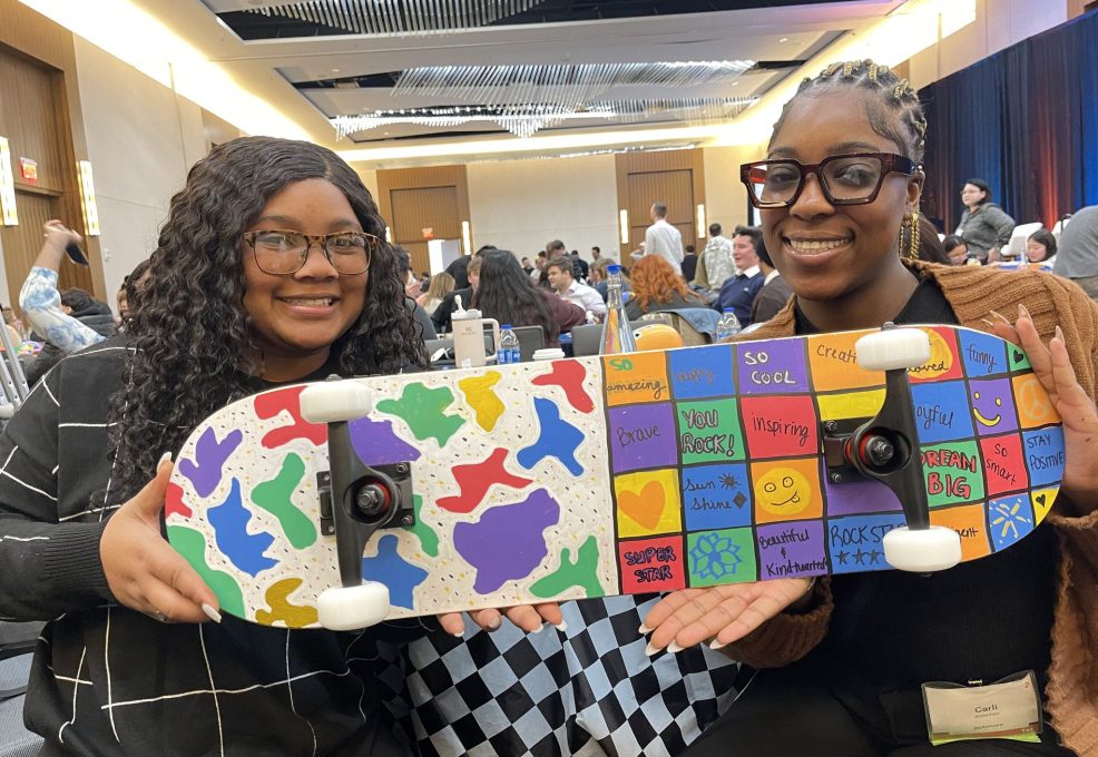 Two participants proudly display their colorful and creatively decorated skateboard, made during the Just Roll With It charitable team building event.