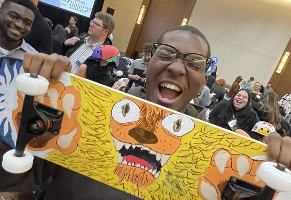 Excited participant proudly holds up a creatively designed skateboard during the Just Roll With It team building event, where teams build and decorate skateboards for charity.