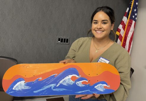 A smiling woman proudly holds up a skateboard that she painted during a team building event with her collages. The design features bright blue waves crashing against an orange and yellow sky, giving the impression of a sunset over the sea.