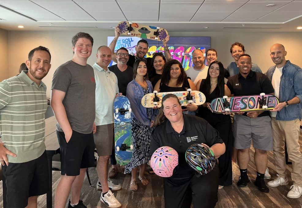 A group of smiling individuals stands together after completing a charitable skateboard build event. They proudly display decorated skateboards and helmets with colorful designs. One skateboard prominently says 'AWESOME' in bold letters, while helmets feature creative, abstract patterns. The group consists of men and women of various ages, all celebrating their accomplishments during this team building event.