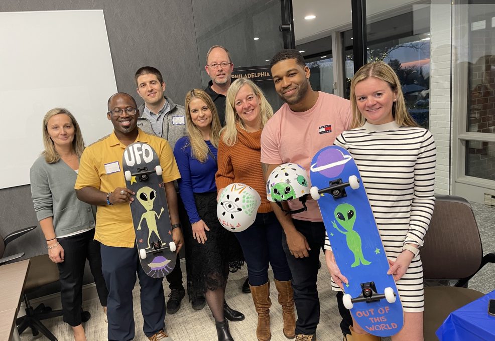 A group of eight people smiling and posing for a picture, holding custom-decorated skateboards and helmets. The skateboards and helmets feature fun designs, including aliens and 'UFO' themes. The group is participating in a team building event where they assemble and decorate skateboards to be donated to charity.