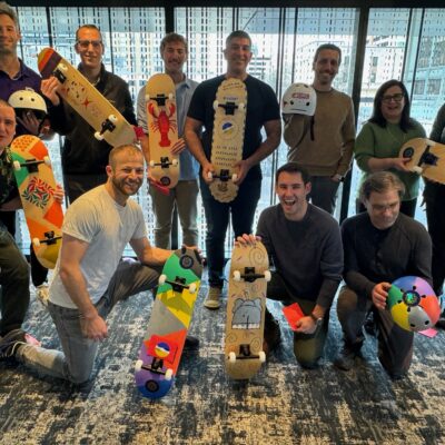 A group of people standing together proudly displaying their custom-designed skateboards and helmets. The skateboards are creatively painted with vibrant colors and designs, showcasing individuality and team spirit. Everyone is smiling, reflecting the success of their collaborative efforts in this charitable team building event. thumbnail
