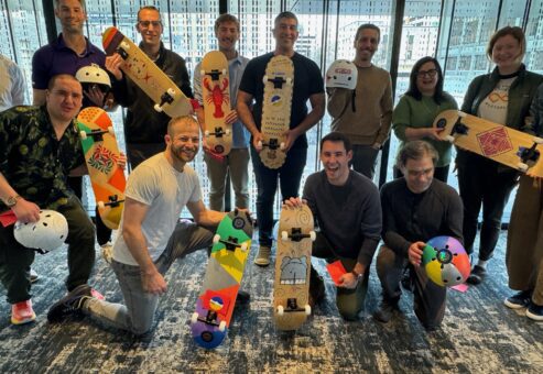 A group of people standing together proudly displaying their custom-designed skateboards and helmets. The skateboards are creatively painted with vibrant colors and designs, showcasing individuality and team spirit. Everyone is smiling, reflecting the success of their collaborative efforts in this charitable team building event.