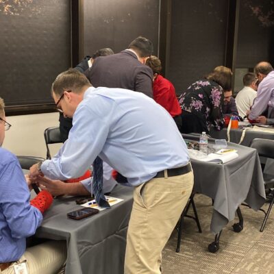 Teams of participants are gathered at tables in a professional setting, focused on assembling prosthetic hands during a team building event. The participants are paired up, wearing red empathy sleeves, and working together on the project. The room has a collaborative atmosphere, with individuals helping each other as they handle tools and components. thumbnail