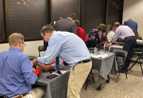 Teams of participants are gathered at tables in a professional setting, focused on assembling prosthetic hands during a team building event. The participants are paired up, wearing red empathy sleeves, and working together on the project. The room has a collaborative atmosphere, with individuals helping each other as they handle tools and components.