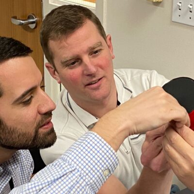 Two men collaborate on assembling a prosthetic hand. Both are focused and working closely together, one man holds a part of the prosthetic while the other assists in making adjustments. The atmosphere is one of teamwork and precision as they carefully work to ensure the hand is correctly assembled. This activity is part of a team building exercise aimed at making prosthetics for those in need. thumbnail