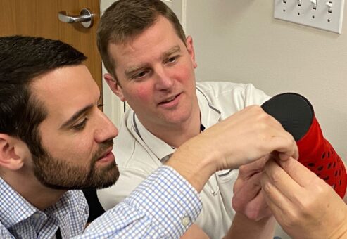 Two men collaborate on assembling a prosthetic hand. Both are focused and working closely together, one man holds a part of the prosthetic while the other assists in making adjustments. The atmosphere is one of teamwork and precision as they carefully work to ensure the hand is correctly assembled. This activity is part of a team building exercise aimed at making prosthetics for those in need.