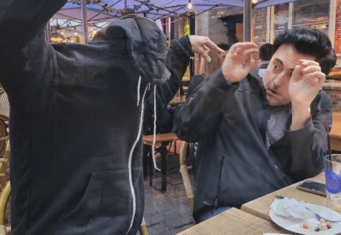 Two people sitting at an outdoor table appear to be acting out a playful, dramatic scene. The person on the left is wearing a hoodie with the hood pulled over their face, raising their arms as if to 'scare' the other person. The person on the right is recoiling in mock fear, raising their hands defensively while leaning back in their chair. The scene takes place under a canopy with string lights, and there are empty plates on the table, suggesting the duo is at a restaurant or cafe.