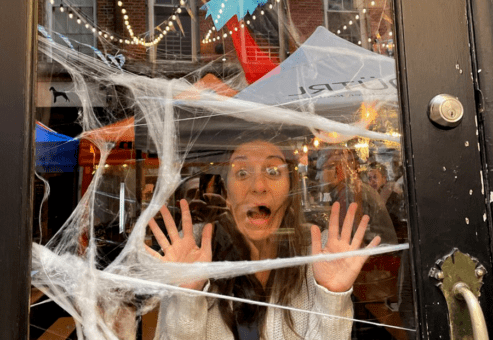 A woman stands behind a glass door decorated with fake spider webs, pressing her hands and face against the glass in a playful expression of fear, as though she's trapped. Behind her, festive lights are strung across an outdoor space, and a red-and-black decorative figure hangs above, adding to the Halloween atmosphere.