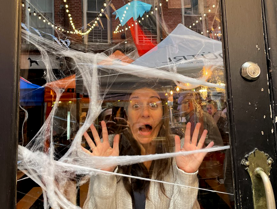 A woman stands behind a glass door decorated with fake spider webs, pressing her hands and face against the glass in a playful expression of fear, as though she's trapped. Behind her, festive lights are strung across an outdoor space, and a red-and-black decorative figure hangs above, adding to the Halloween atmosphere.
