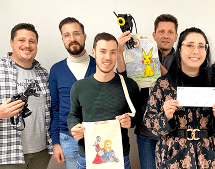 A group of five people stand together, proudly holding items they have worked on during the team building: Prosthetic Hand Project. One person holds a prosthetic hand, and others display creatively decorated hand-storage bags featuring colorful designs, such as Pikachu and a superhero. The group is smiling, demonstrating teamwork and creativity, while contributing to an initiative aimed at helping amputees in developing countries regain functionality and independence.