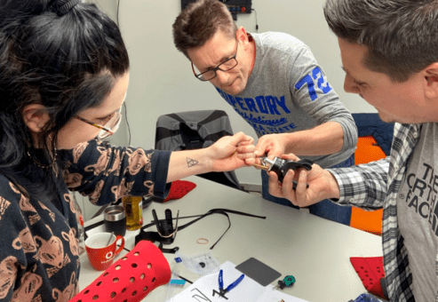 Three individuals are actively collaborating on assembling a prosthetic hand as part of The Prosthetic Hand Project team building activity. One person holds a component while another uses a tool to adjust part of the prosthetic. The third person watches and assists in the process. Various tools, materials, and instructions are spread across the table, including a hand-storage bag, reinforcing the creative and practical nature of the task.