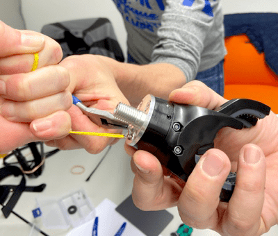 A close-up of two individuals' hands working together to assemble the mechanical claw of a prosthetic hand during The Prosthetic Hand Project. One person is holding the claw component while the other uses a tool to fasten or adjust a yellow cord. The detailed view showcases the precision and teamwork required for the assembly process.