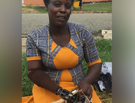 A woman sitting outdoors smiles while using a prosthetic hand to weave a basket. She wears a colorful orange and blue dress, and her activity highlights the practical benefits of the prosthetic, allowing her to engage in skilled handiwork. This image demonstrates how the Prosthetic Hand Project supports amputees in developing countries by enabling them to perform everyday tasks and regain their independence.