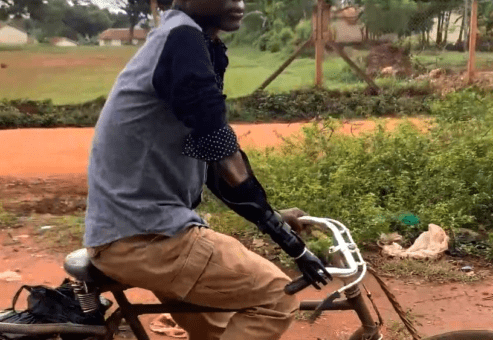 A man rides a bicycle along a dirt path in a rural area. He is wearing a long-sleeved shirt and pants, with a prosthetic arm visible on his left side, gripping the handlebar. The man appears focused, demonstrating how the prosthetic arm allows him to engage in everyday activities.