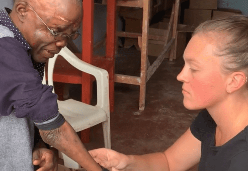 A woman carefully fits a prosthetic arm on an older man in a workshop setting. The man, sitting on a plastic chair, looks down at the prosthetic as the woman adjusts it, focused on ensuring a proper fit. The man wears glasses and a long-sleeved shirt, with his new prosthetic visible on his left arm. This moment captures the emotional and practical significance of receiving a prosthetic, a life-changing experience for the man.