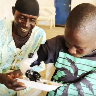 A young boy with a prosthetic hand excitedly uses it to eat from a plate, while an older man, smiling warmly, assists him. The scene captures a moment of joy and empowerment, highlighting the transformative impact of the Prosthetic Hand Project in improving daily life for individuals. thumbnail