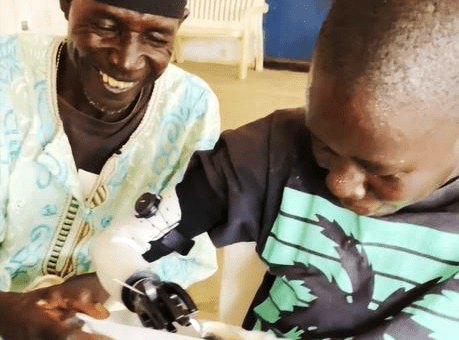A young boy with a prosthetic hand excitedly uses it to eat from a plate, while an older man, smiling warmly, assists him. The scene captures a moment of joy and empowerment, highlighting the transformative impact of the Prosthetic Hand Project in improving daily life for individuals.