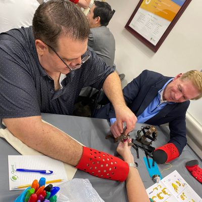 Two participants are actively assembling a prosthetic hand during The Prosthetic Hand Project. They are wearing red prosthetic simulation sleeves, adding a level of challenge and empathy to the task. One person is holding and connecting the components of the mechanical hand while the other assists. A variety of tools and assembly instructions are visible on the table, and a notebook and colorful pens are seen nearby, indicating careful planning and attention to detail. This image highlights the teamwork, focus, and effort involved in assembling prosthetic hands for donation to those in need.