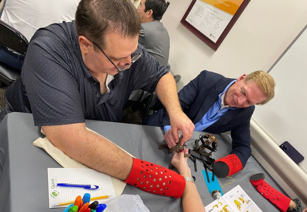 Two participants are actively assembling a prosthetic hand during The Prosthetic Hand Project. They are wearing red prosthetic simulation sleeves, adding a level of challenge and empathy to the task. One person is holding and connecting the components of the mechanical hand while the other assists. A variety of tools and assembly instructions are visible on the table, and a notebook and colorful pens are seen nearby, indicating careful planning and attention to detail. This image highlights the teamwork, focus, and effort involved in assembling prosthetic hands for donation to those in need.
