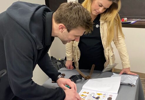 Two individuals are focused on assembling a prosthetic hand as part of The Prosthetic Hand Project. One person is intently working with components from a small plastic case, following the provided instructions laid out on the table. The second individual looks on, offering support and guidance during the assembly process. The setup on the table includes detailed assembly guides and various parts for the hand. This image captures teamwork, concentration, and the hands-on effort involved in building prosthetic hands during a team building program.