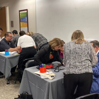 A group of individuals is seated at tables working on a team building activity in a conference room. Each table has participants intently focused on assembling prosthetic hands as part of a charitable event. Participants are wearing red empathy sleeves on their arms while carefully working with tools and parts. thumbnail