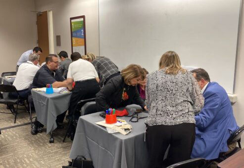 A group of individuals is seated at tables working on a team building activity in a conference room. Each table has participants intently focused on assembling prosthetic hands as part of a charitable event. Participants are wearing red empathy sleeves on their arms while carefully working with tools and parts.