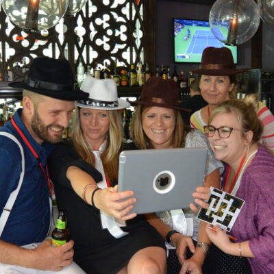 Team members wearing detective hats gathered around a tablet during the Sleuth: The Murder Mystery Scavenger Hunt team building activity, smiling and analyzing clues together thumbnail