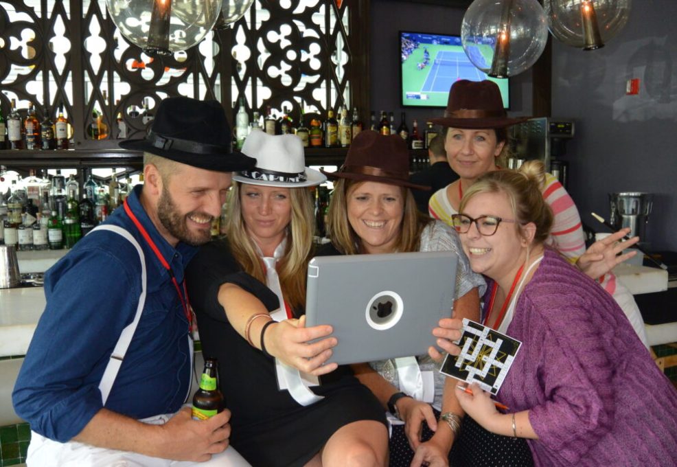 Team members wearing detective hats gathered around a tablet during the Sleuth: The Murder Mystery Scavenger Hunt team building activity, smiling and analyzing clues together