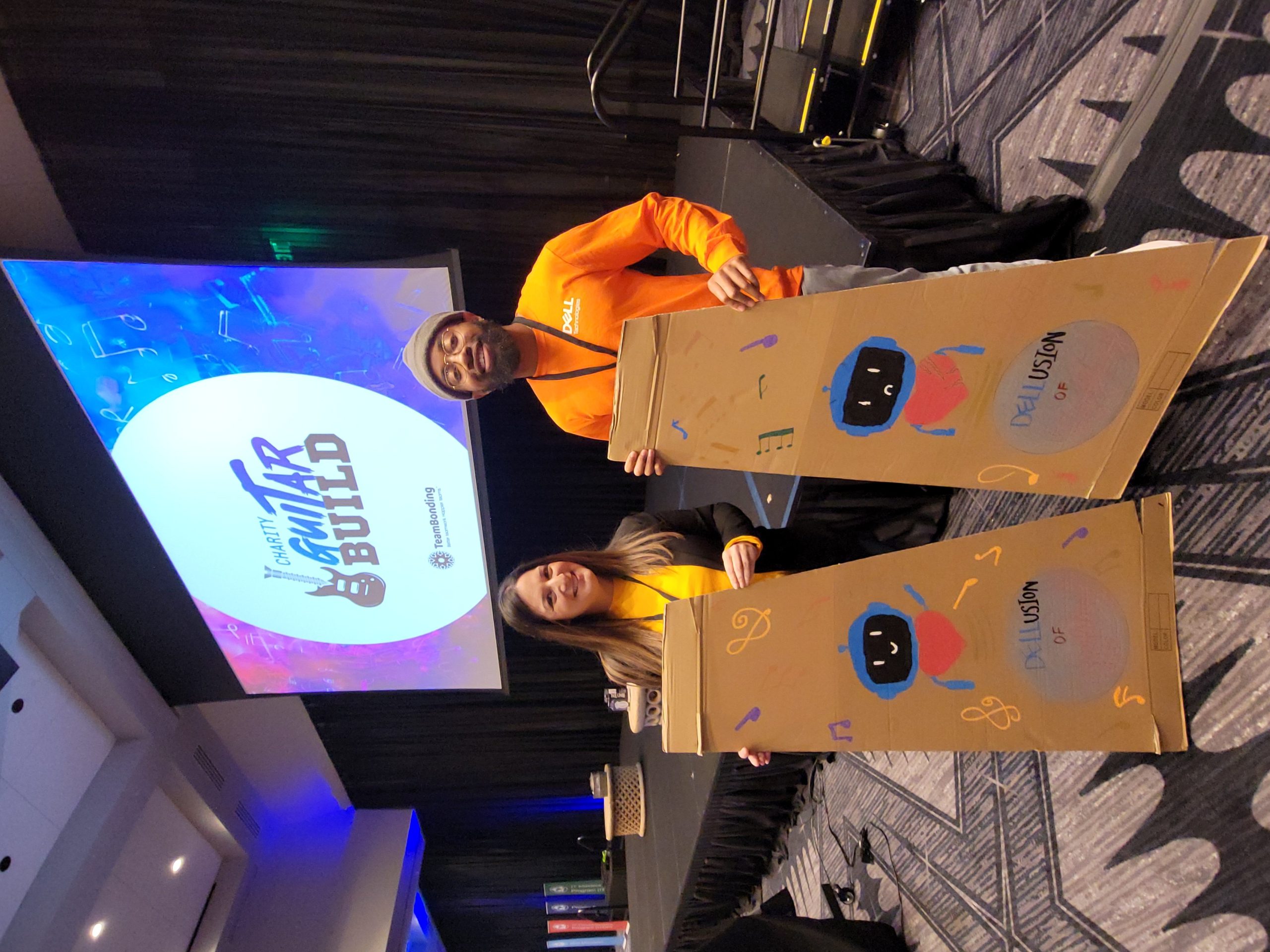 Two team members proudly display their decorated cardboard guitar cases during the Charity Guitar Build team building event, showcasing their creativity and enthusiasm.