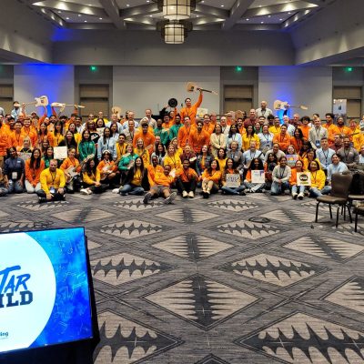 Large group photo at the Charity Guitar Build team building event, with participants holding guitars and celebrating teamwork and community impact. thumbnail