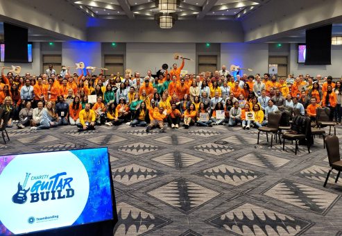 Large group photo at the Charity Guitar Build team building event, with participants holding guitars and celebrating teamwork and community impact.
