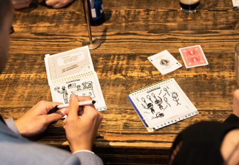 Team building participants engage in a creative drawing game during a business networking event, using cards and sketchpads to spark fun conversations and connections.