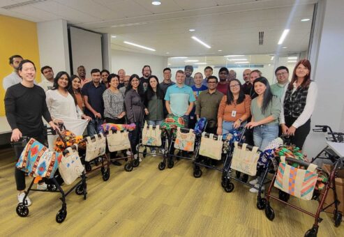 Group of participants posing with completed rollator walkers decorated with colorful blankets and tote bags, smiling after the successful assembly during a charitable team building program.