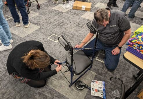 Two participants are kneeling on the floor, carefully assembling a rollator walker by attaching its handles and frame components during a charitable team building activity.